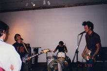 Load image into Gallery viewer, A band performing live on stage June 24th, 1999, with a guitarist in the foreground, a drummer behind, and bass player near a keyboard, captured in a tilted, low-light photograph.