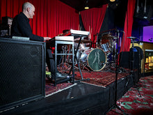 Load image into Gallery viewer, Musician playing keyboard on stage with drum set, red curtains, and patterned carpet.