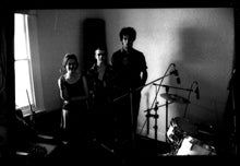 Load image into Gallery viewer, Black and white photograph of a band performing in a room, with a drummer, bassist, and vocalist visible in a dimly lit interior with a window.